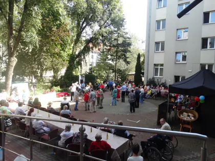 Piknik na podwórku Domu, goście tańczą i siedzą przy długich stołach w cieniu drzew