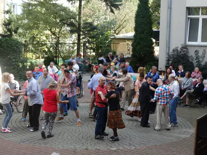 Pary tańczą na wybrukowanym podwórku, wokół siedzą goście, w tle kamienica i drzewa