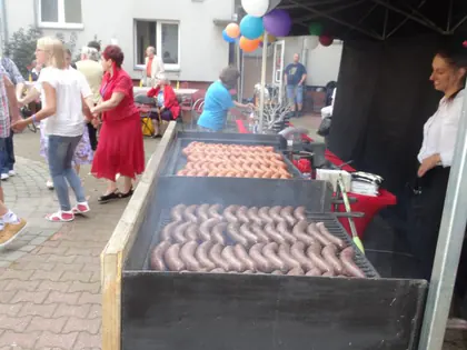 Grill z kiełbaskami pod czarnym namiotem, w tle goście pikniku i balony