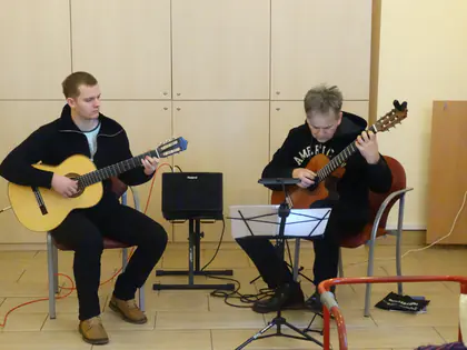 Dwóch mężczyzn gra na gitarach klasycznych, między nimi pulpit z nutami i wzmacniacz Roland