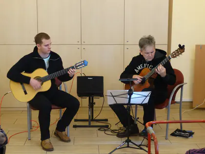 Dwóch mężczyzn gra na gitarach klasycznych, przed nimi stoi pulpit z nutami i wzmacniacz