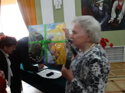 Starsza kobieta z mikrofonem trzyma prezent w zielonej wstążce w sali ze zdobionymi ścianami