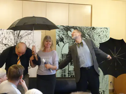 Troje aktorów z parasolami odgrywa scenkę przed dekoracją z kamienicą