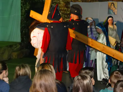 Żołnierze w czerwonych strojach prowadzą Jezusa z krzyżem przed widzami na sali gimnastycznej