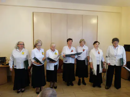 Siedem pań z chóru w białych bluzkach i czarnych spódnicach śpiewa z nutami w dłoniach