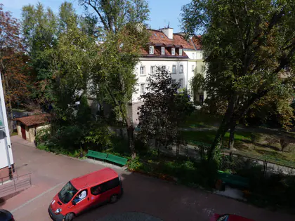 Widok z okna na zielony ogród, kamienicę i czerwony bus zaparkowany na podwórku