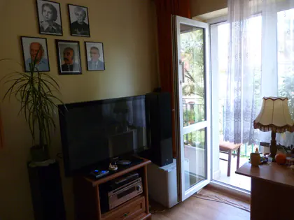 Pokój mieszkańca z telewizorem, lampką i drzwiami balkonowymi, na ścianie oprawione portrety