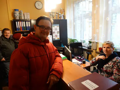Mieszkaniec w czerwonej kurtce stoi przy biurku, obok siedzi pracownica biura