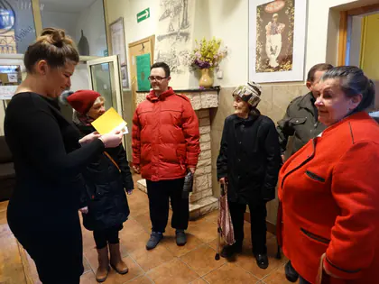 Kobieta czyta kartkę mieszkańcom ubranym do wyjścia w holu Domu