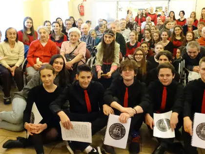 Chór licealny w czerwonych krawatach siedzi wśród mieszkańców w świetlicy