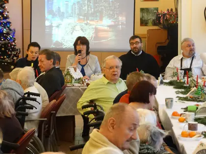 Mieszkańcy przy wigilijnym stole, kobieta przemawia do mikrofonu, obok zakonnik