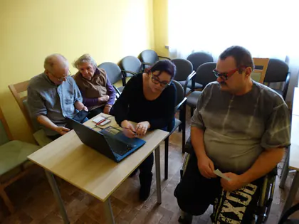 Mieszkańcy siedzą przy stoliku z laptopem, kobieta pokazuje im coś na ekranie