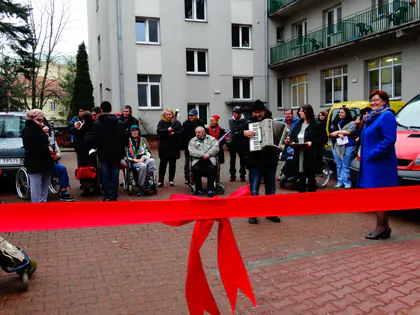 Mieszkańcy, goście i muzyk z akordeonem przy czerwonej wstędze przed budynkiem Domu