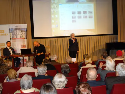 Prelegentka mówi do mikrofonu na scenie przed publicznością seniorów