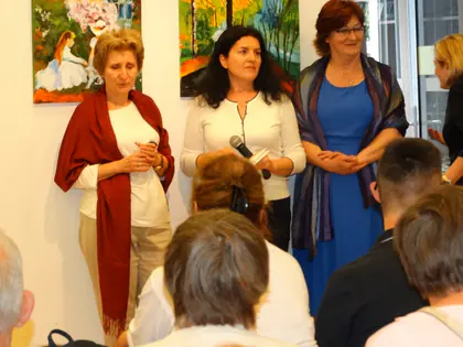 Trzy kobiety, jedna z mikrofonem, stoją przed publicznością podczas wernisażu