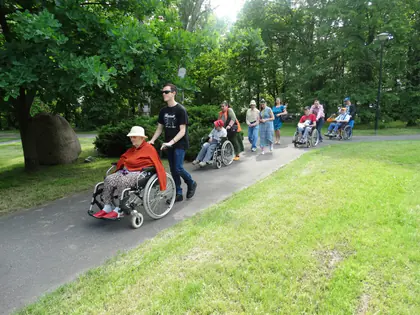 Opiekunowie i mieszkańcy na wózkach jadą długą alejką przez zielony park