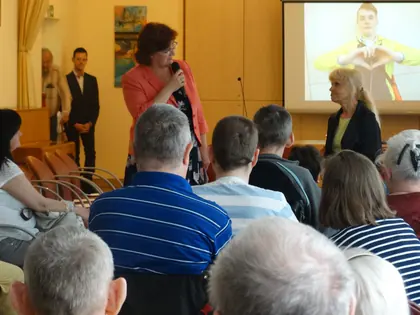 Prowadząca mówi do mikrofonu przed widownią, na ekranie postać pokazująca serce dłońmi