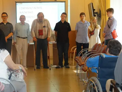 Młodzież i mieszkańcy stoją w szeregu podczas spektaklu przed ekranem z napisami