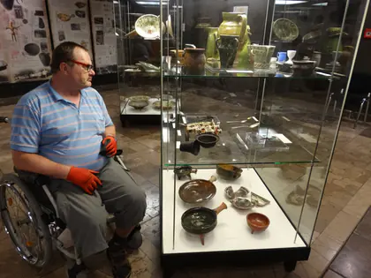 Mieszkaniec na wózku ogląda gablotę z zabytkowymi naczyniami w muzeum