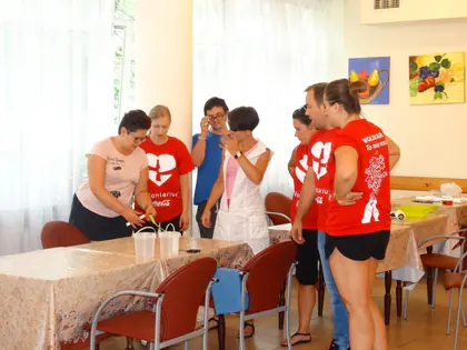 Wolontariusze w czerwonych koszulkach przygotowują coś przy stole razem z opiekunkami
