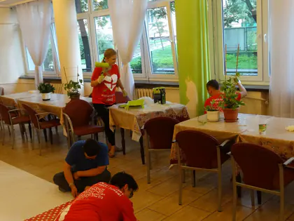 Wolontariusze w czerwonych koszulkach sprzątają i ustawiają kwiaty w jadalni