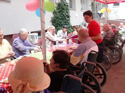Mieszkańcy na wózkach siedzą przy stołach w cieniu namiotów i balonów