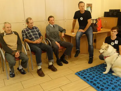 Mieszkańcy siedzą w kręgu, przed nimi biały pies husky na niebieskim kocyku