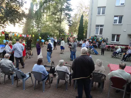 Festyn na podwórku Domu, goście siedzą przy stołach i spacerują wśród kolorowych balonów