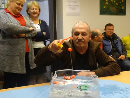 Mieszkaniec łowi wędką zabawkowe rybki z akwarium na stole, obok przyglądają się dwie panie