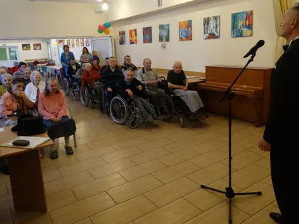 Piosenkarz w smokingu występuje przed mieszkańcami siedzącymi na krzesłach i wózkach