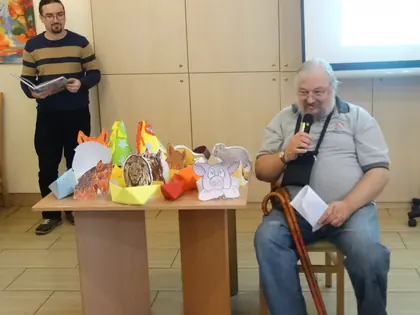 Mieszkaniec z laską czyta bajkę do mikrofonu, obok stół z papierowymi figurkami