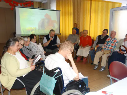 Mieszkańcy siedzą w kręgu, mężczyzna czyta przez mikrofon, w tle ekran z wielkanocnym obrazem