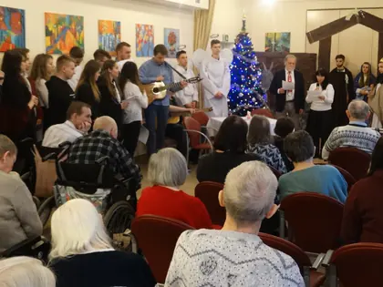 Młodzież śpiewa kolędy z gitarą przed mieszkańcami siedzącymi w rzędach