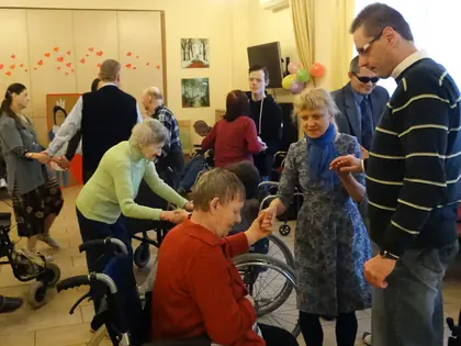 Mieszkanki tańczą z gośćmi, trzymając się za ręce, część osób na wózkach