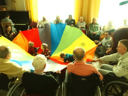 Mieszkańcy na wózkach unoszą kolorową chustę, pod nią bawią się dzieci