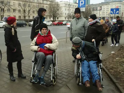 Mieszkańcy na wózkach i opiekunowie na chodniku przy ruchliwej ulicy