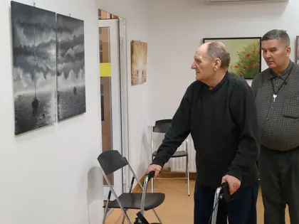 Mieszkaniec z balkonikiem ogląda obrazy, obok drugi mężczyzna