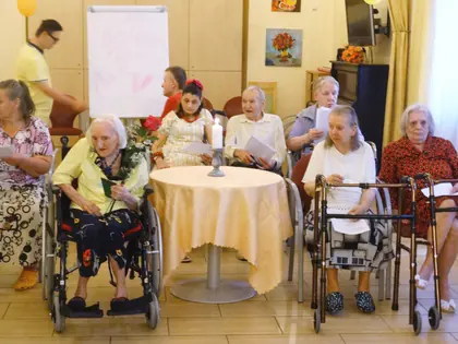 Jubilatka na wózku z kwiatami wśród mieszkańców przy stole ze świecą