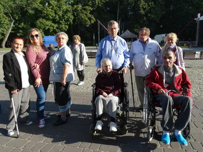 Grupa mieszkańców i opiekunów stoi na placu, dwoje na wózkach