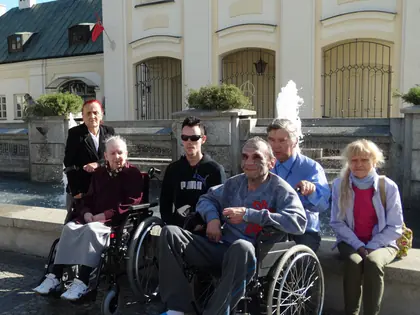 Mieszkańcy na wózkach i opiekunowie pozują przed zabytkowym budynkiem z fontanną