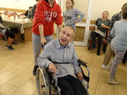 Opiekunka pcha wózek z uśmiechniętym mieszkańcem przez świetlicę