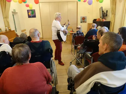Gitarzystka śpiewa przechodząc między mieszkańcami siedzącymi w świetlicy