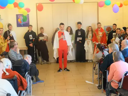 Chłopak w czerwonym stroju mówi do mikrofonu, wokół młodzież w kostiumach świętych
