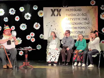Aktorzy siedzą na scenie przed banerem przeglądu, jeden w kolorowym kapeluszu