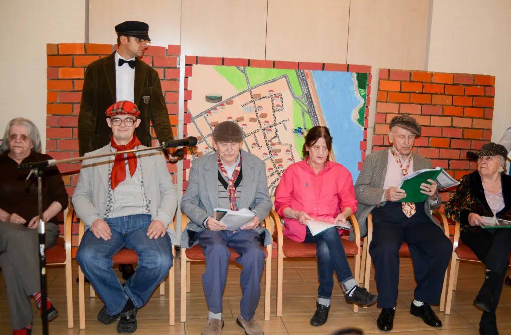 Mieszkańcy w kostiumach siedzą na scenie przed dekoracją z cegieł i planem miasta.