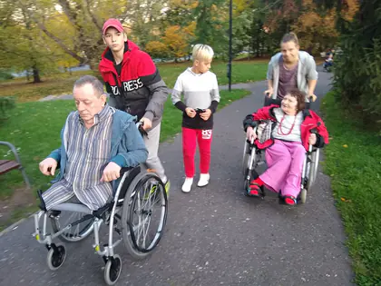 Młodzież pcha wózki z mieszkańcami parkową alejką