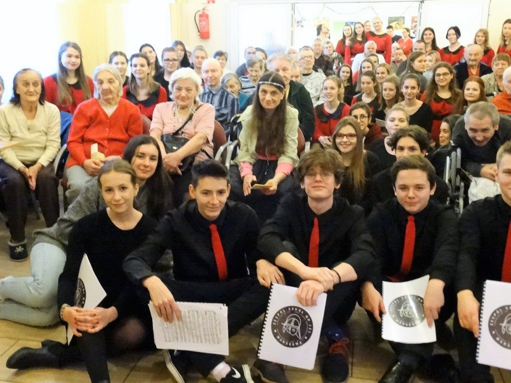 Mieszkańcy i młodzież w czerwono-czarnych strojach na wspólnym koncercie w świetlicy.