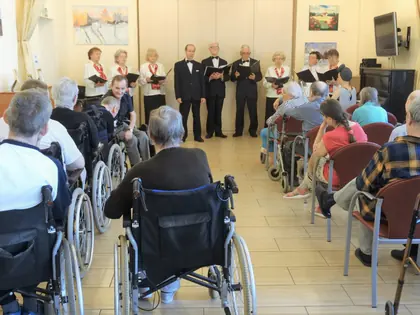 Mieszkańcy na wózkach słuchają występu chóru w świetlicy