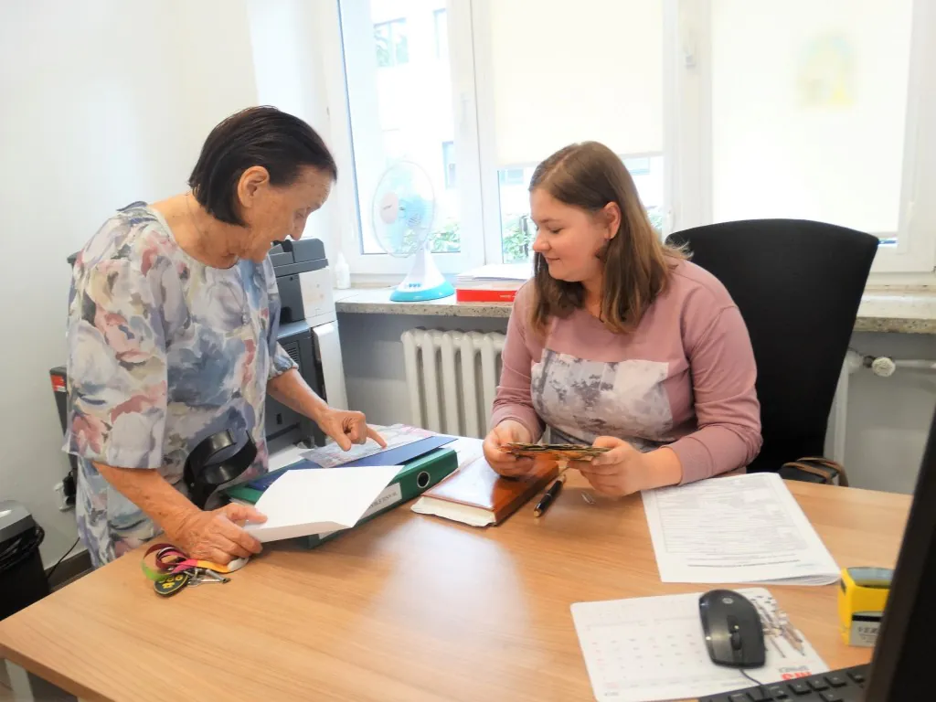 Pracownica socjalna przy biurku rozmawia z mieszkanką, razem przeglądają dokumenty.