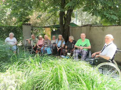 Mieszkańcy na wózkach i ławkach odpoczywają w ogrodzie za trawami